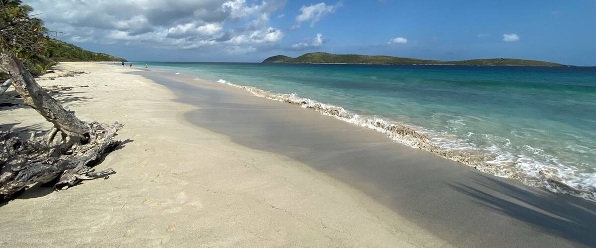A beach with clear turquoise water, a sandy shore, and distant green hills under a partly cloudy sky.