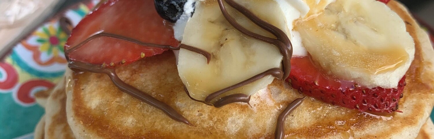 Uma pilha de panquecas cobertas com chantilly, calda de chocolate, morangos, mirtilos e fatias de banana.