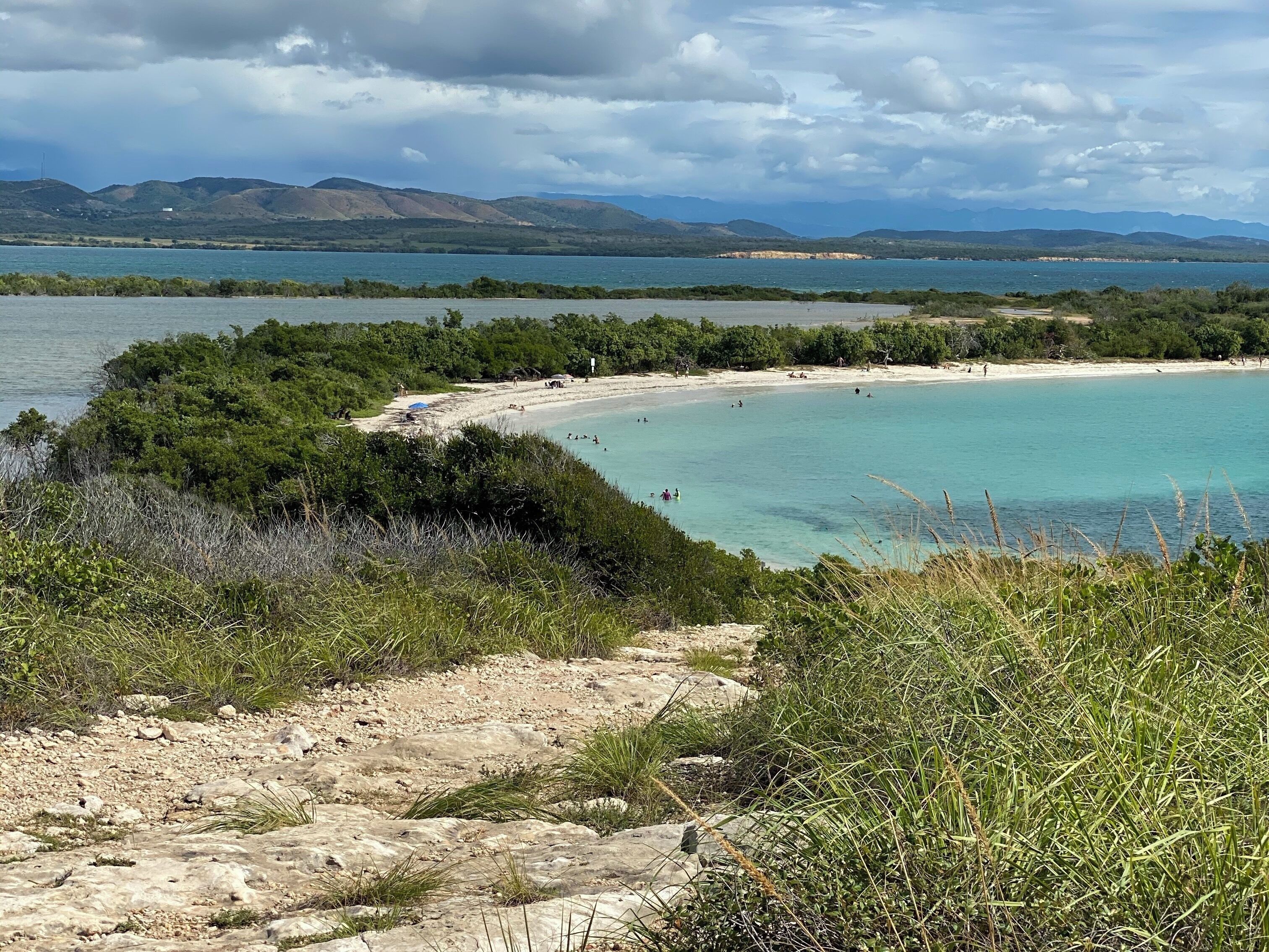A coastal landscape with a sandy beach, clear turquoise waters, and distant mountains under a cloudy sky.