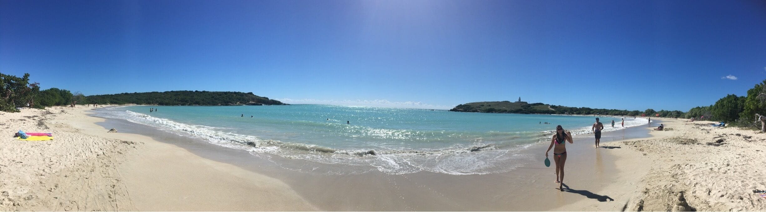 Una playa arenosa con agua turquesa cristalina, algunas personas y colinas al fondo.