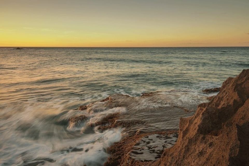 Un paysage côtier avec des vagues qui s’écrasent contre des rivages rocheux, et un coucher de soleil en arrière-plan.