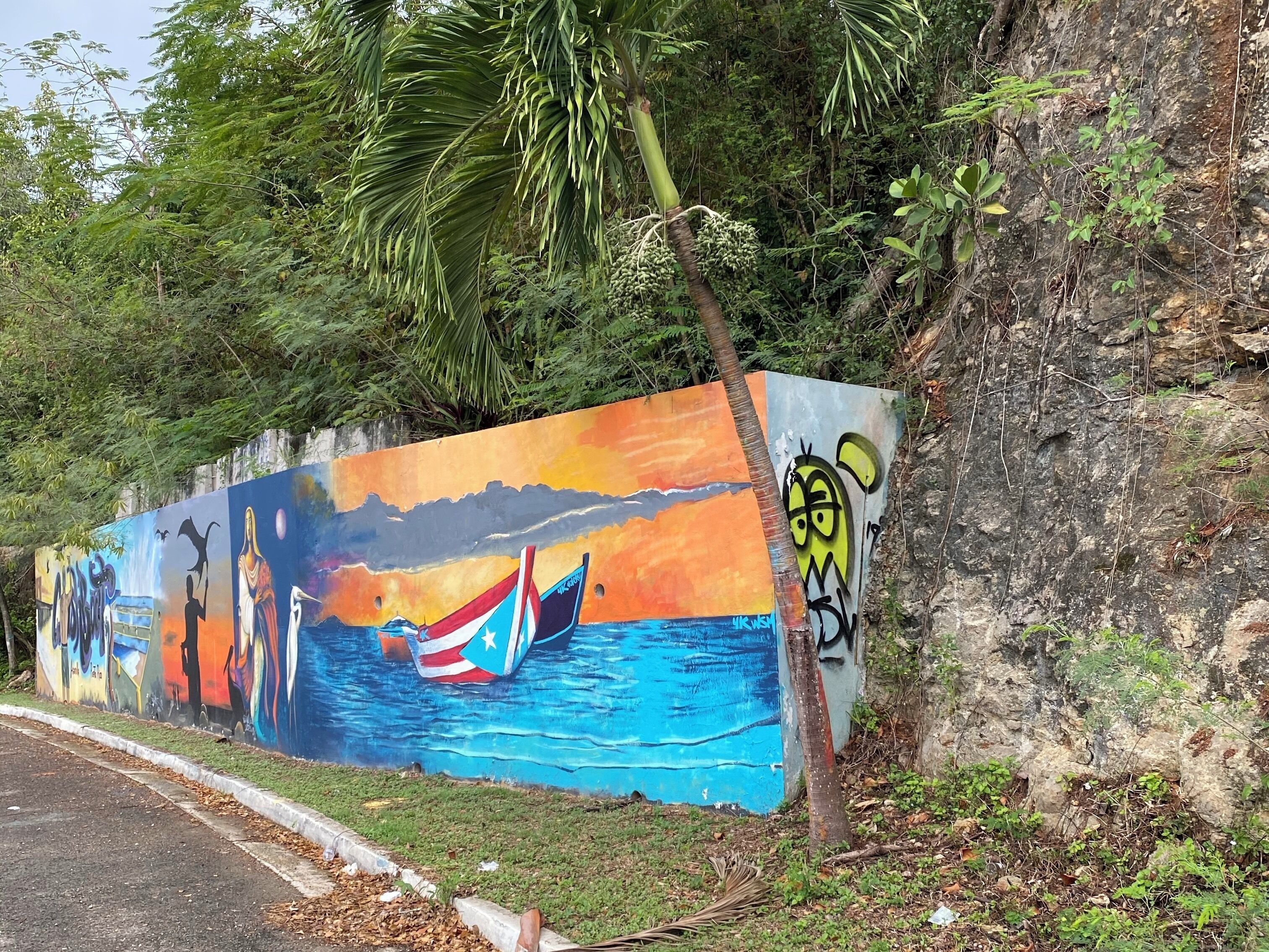 Un mural colorido que muestra un barco, un atardecer y una persona sobre un acantilado.