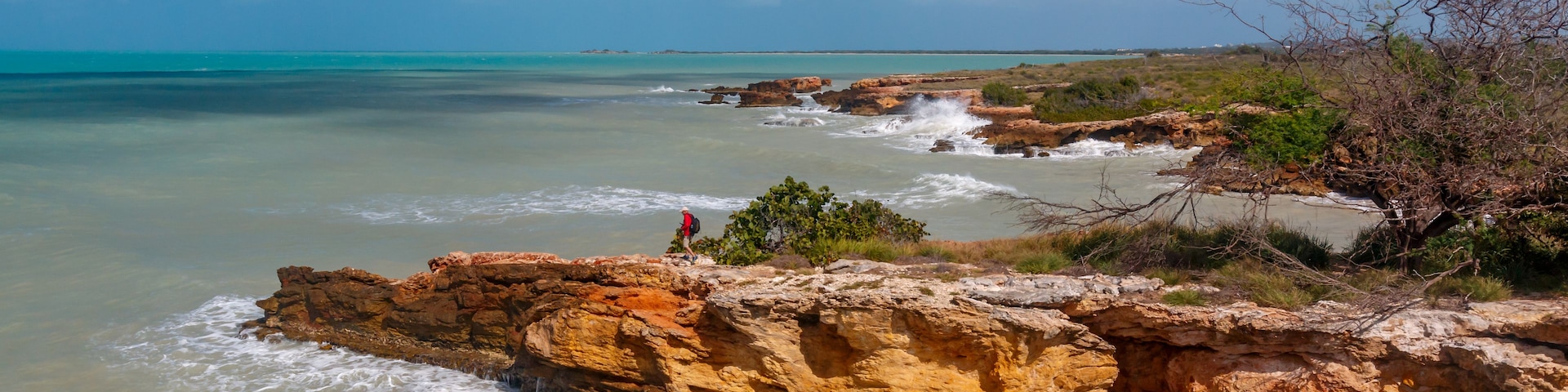 Paisaje costero con acantilados rocosos, aguas turquesas cristalinas y un excursionista.