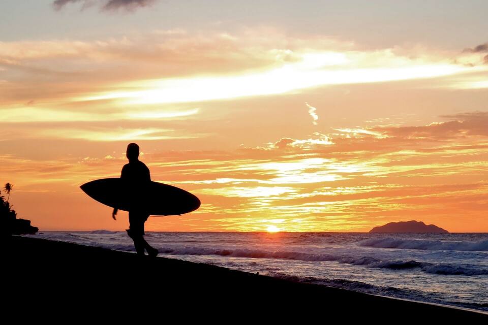 A silhouette of a person carrying a surfboard walking along a beach at sunset.