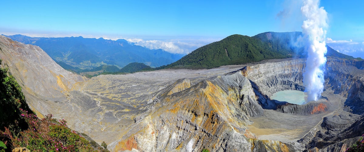A volcanic landscape with a prominent geyser, a crater lake, and surrounding mountains.