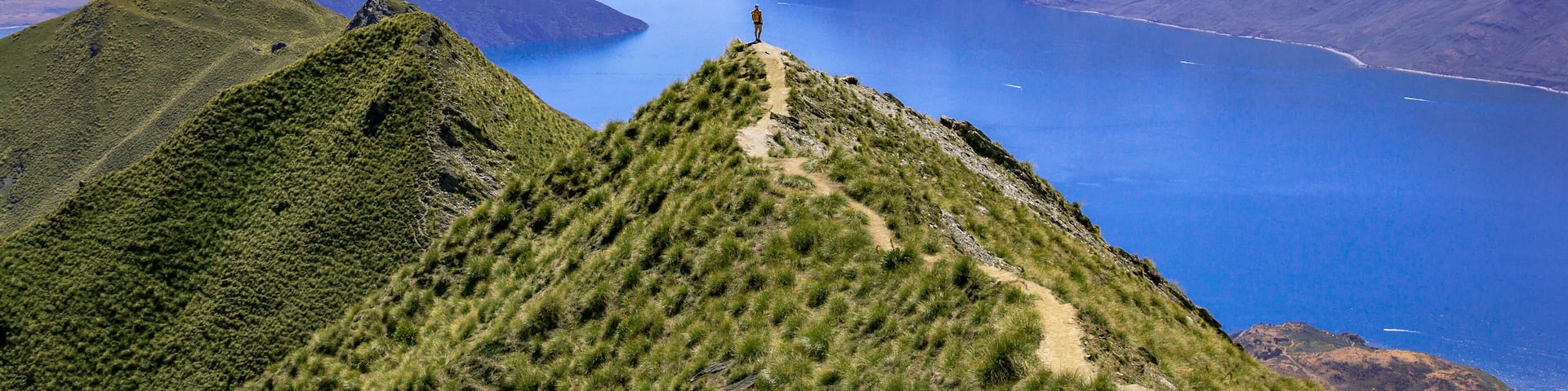 Un excursionista en la cima de una montaña, con vista a un lago extenso rodeado de montañas escarpadas.