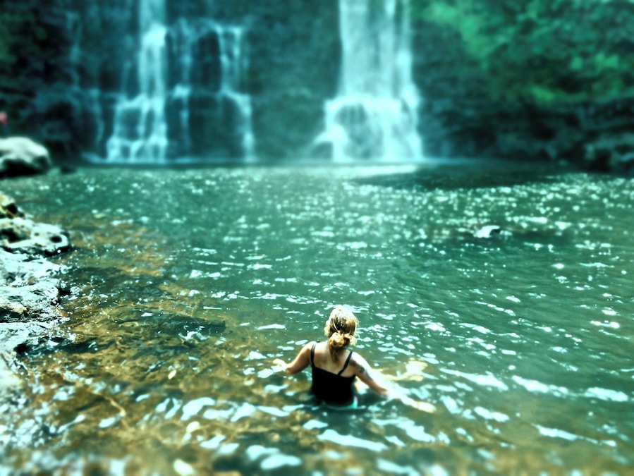 A woman in a black swimsuit standing in clear water with a waterfall in the background.