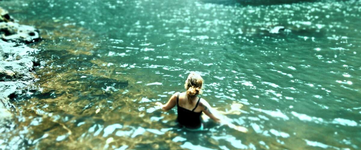 A woman in a black swimsuit standing in clear water with a waterfall in the background.