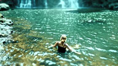 A woman in a black swimsuit standing in a clear river with a waterfall in the background.
