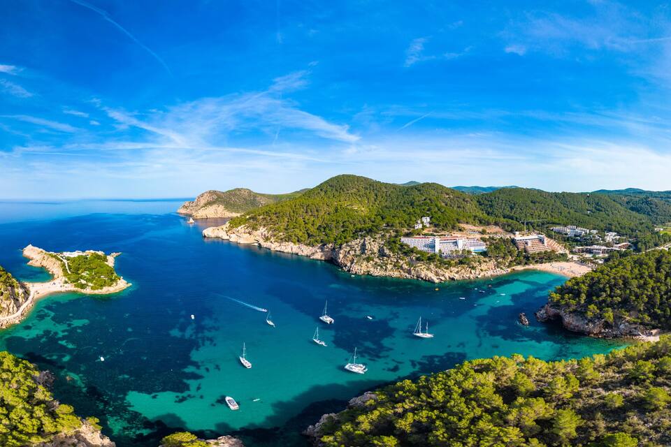 A coastal landscape with clear turquoise waters, a small island with buildings, and surrounding green hills.