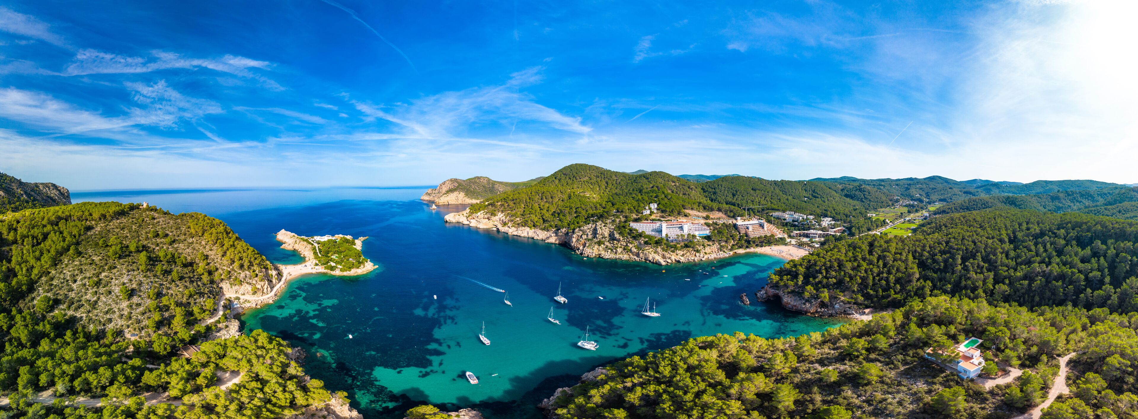 A coastal landscape with clear turquoise waters, a small island with buildings, and surrounding green hills.