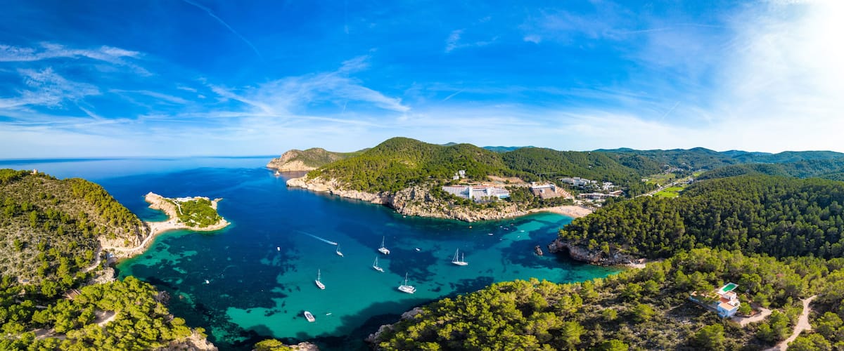 A coastal landscape with clear turquoise waters, a small island with buildings, and surrounding green hills.