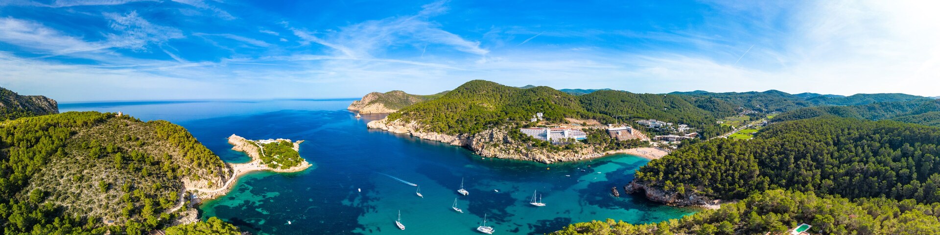 A coastal landscape with clear turquoise waters, a small island with buildings, and surrounding green hills.