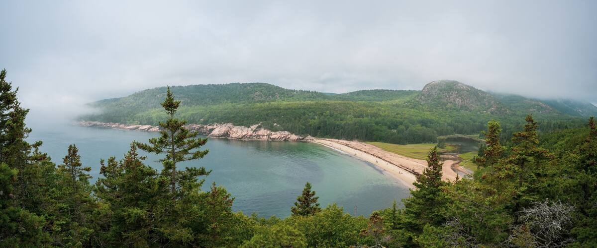 A coastal landscape with a sandy beach, dense forest, and a bay surrounded by hills.