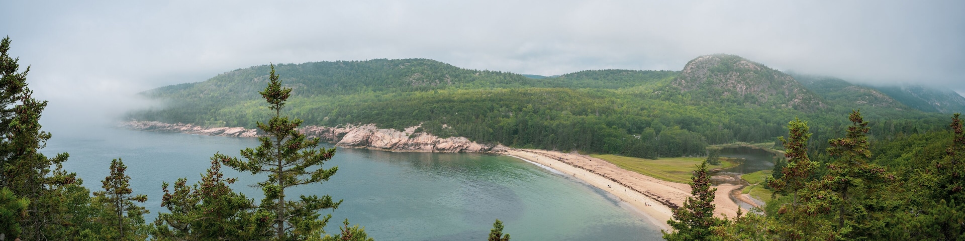 A coastal landscape with a sandy beach, dense forest, and a bay surrounded by hills.