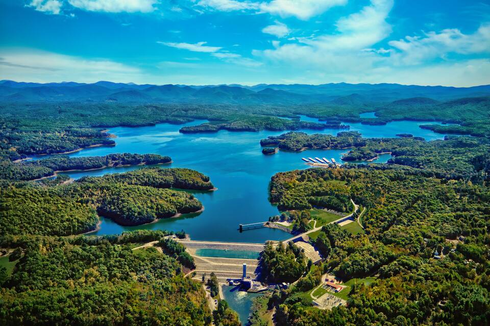 A large lake surrounded by forested hills, a dam, and a small dock.