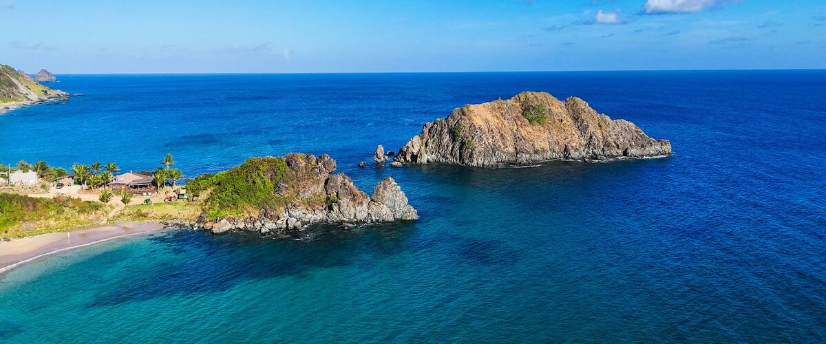 A coastal landscape with clear blue waters, rocky islands, and a small settlement with buildings and palm trees.