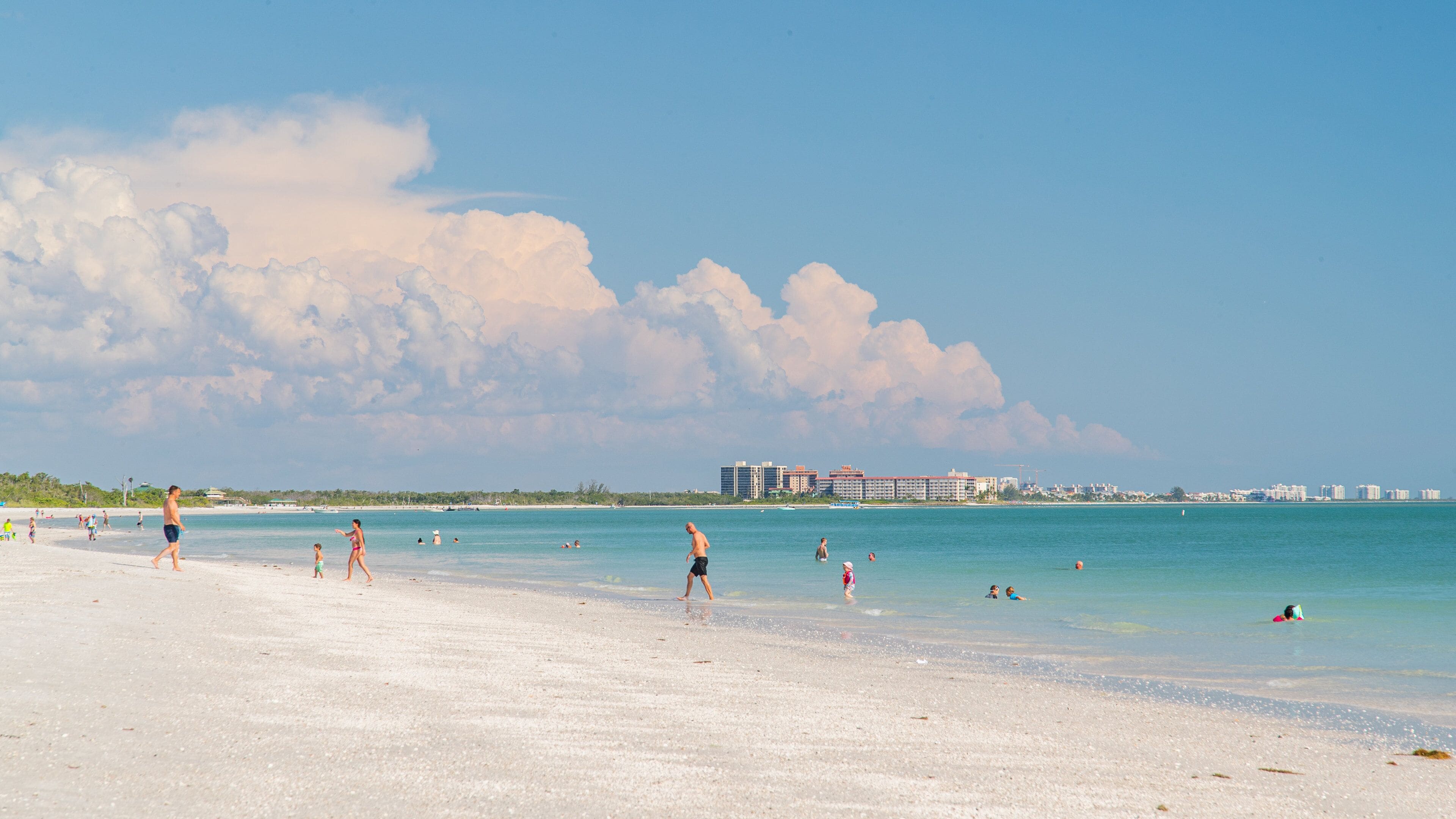 A beach with people walking and swimming, a clear sky, and buildings in the background.