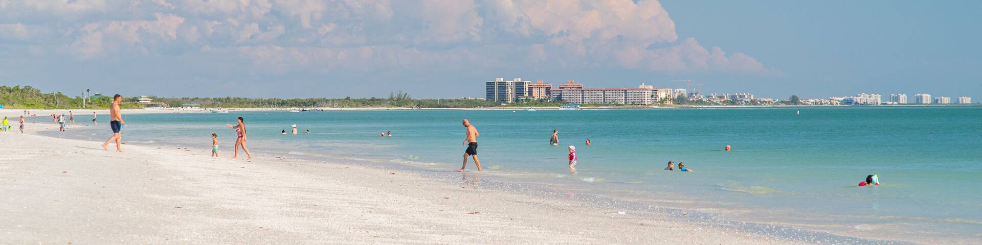 A beach with people walking and swimming, a clear sky, and buildings in the background.