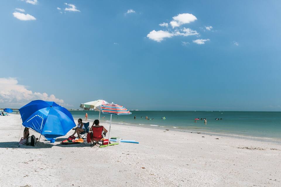 Uma praia com pessoas relaxando sob guarda-sóis, céu azul límpido e águas calmas do oceano.