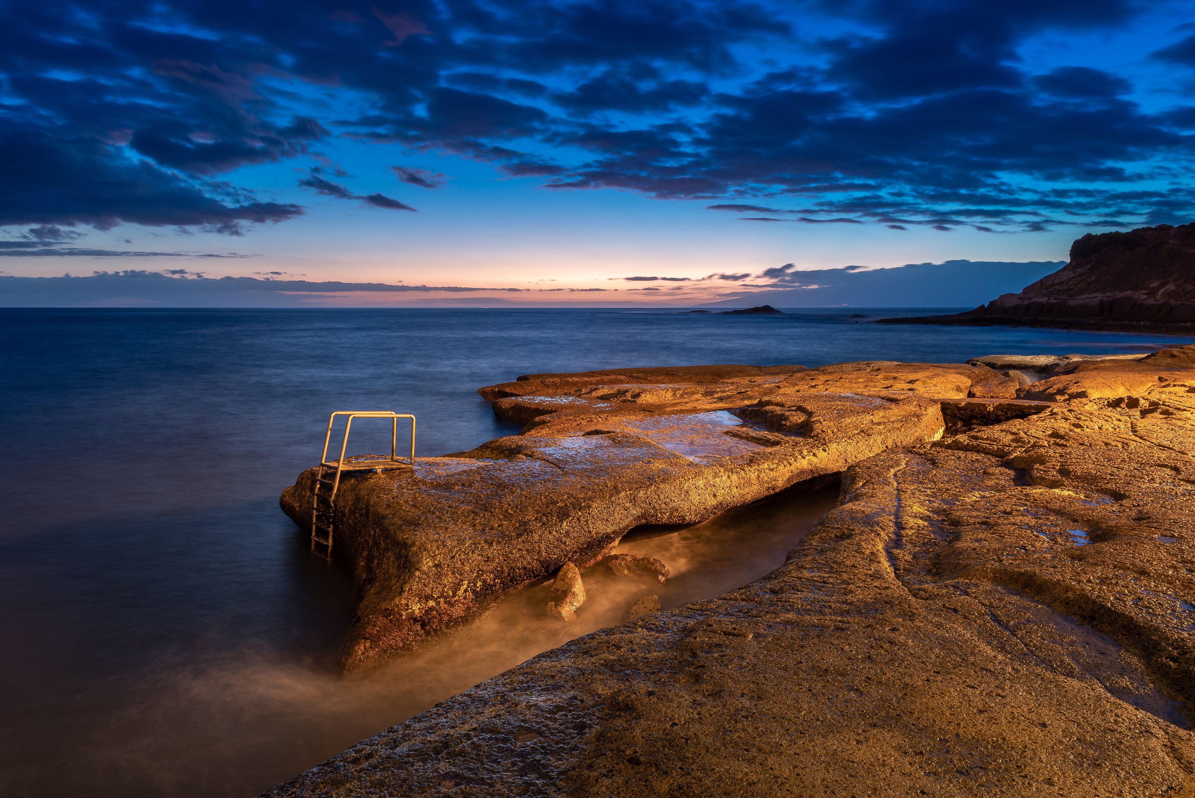 Costa rocosa al atardecer con una escalera sobre una formación rocosa.