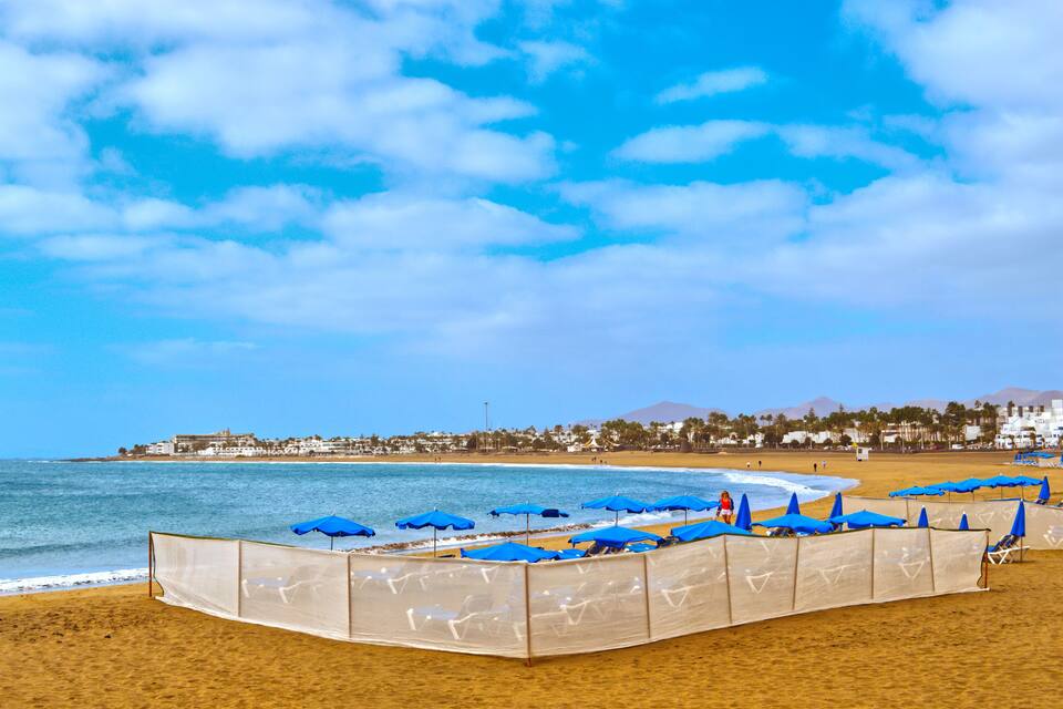 Una playa con sombrillas azules y tumbonas, un cielo azul despejado y una cordillera distante.