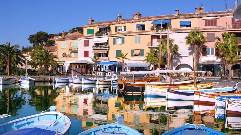 A marina with colorful boats docked, surrounded by Mediterranean-style buildings and palm trees.