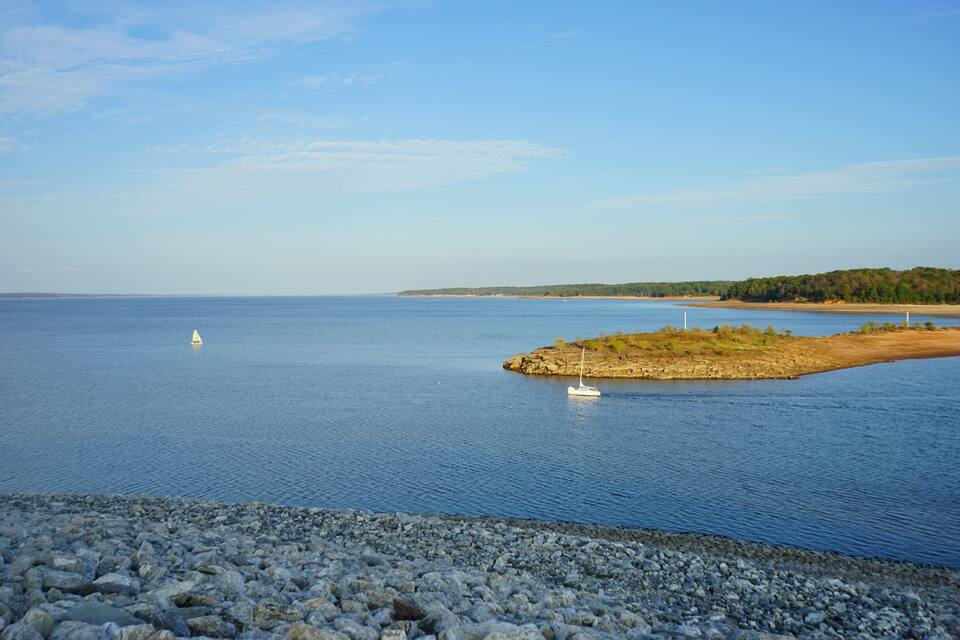 A calm lake with a rocky shoreline, a small island with sparse vegetation, and a sailboat in the distance.