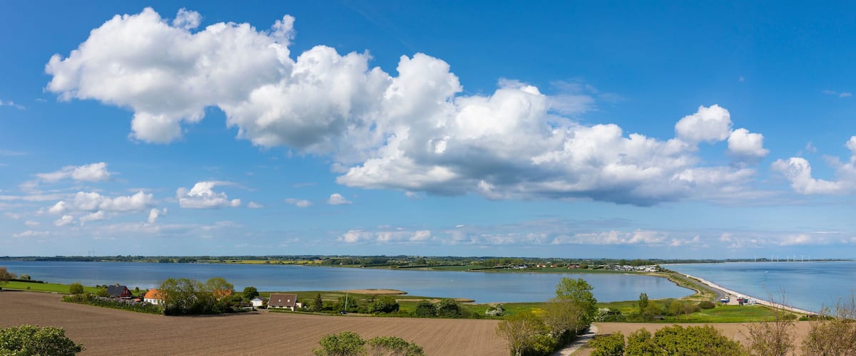 A coastal landscape with a large body of water, fields, and a distant shoreline with wind turbines.