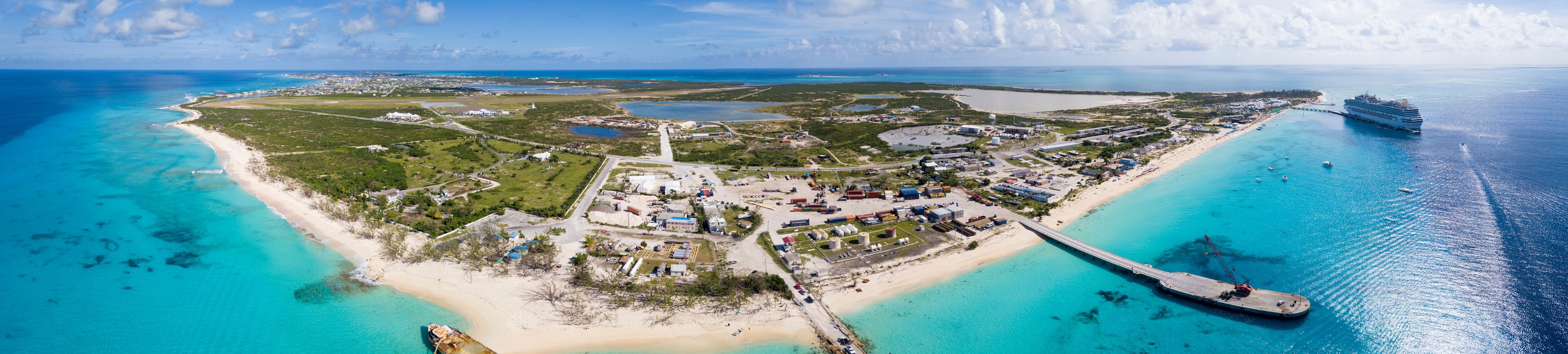 Vue aérienne d’une zone côtière où l’on aperçoit un grand navire de croisière amarré, une jetée s’étendant dans les eaux turquoise, ainsi qu’un mélange de bâtiments industriels et résidentiels.