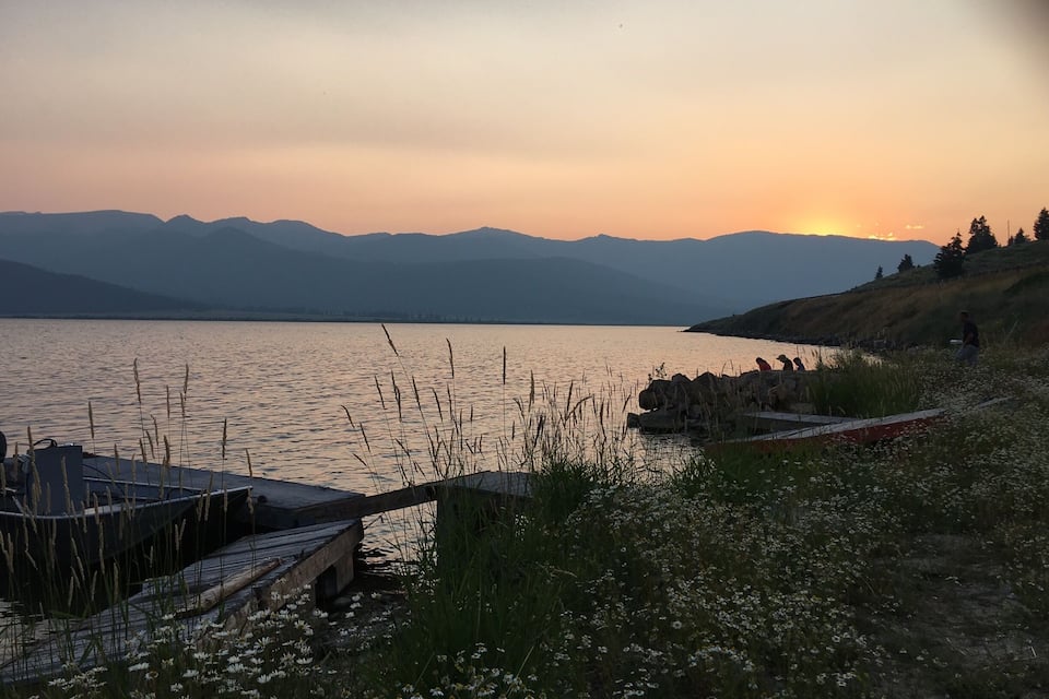 Ein Sonnenuntergang am Seeufer mit Bergen im Hintergrund und einer hölzernen Anlegestelle.