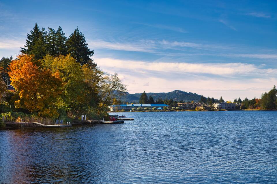Um lago tranquilo com folhagem de outono, um cais e edifícios ao fundo.