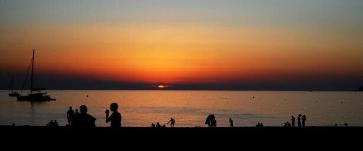 A sunset over a calm sea with silhouettes of people and a sailboat.