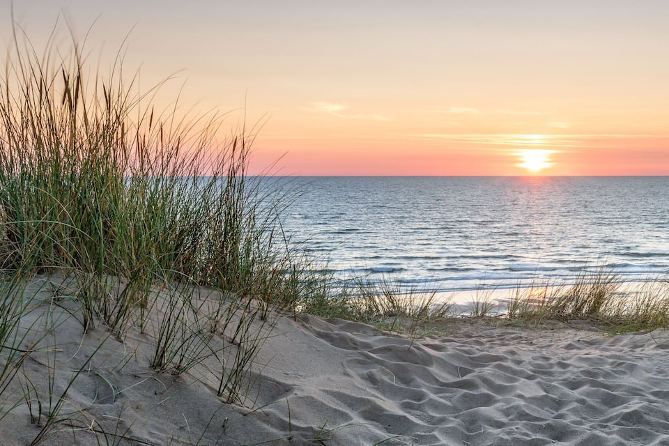 Una playa con dunas de arena y césped en primer plano, que conduce a un mar tranquilo bajo un cielo al atardecer.