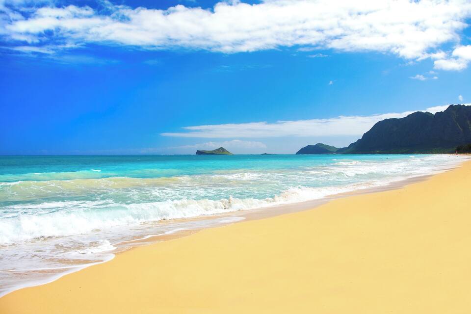 A beach with clear turquoise water, a sandy shore, and distant mountains under a blue sky.