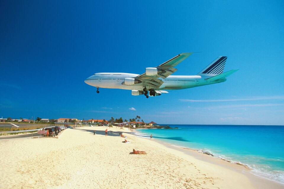 Ett stort flygplan flyger lågt över en strand med klart, blått vatten och en sandstrand.