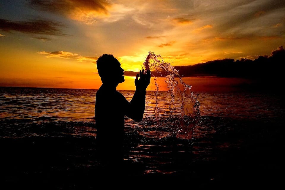 A silhouette of a person playing with water against a sunset over the ocean.