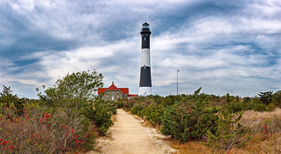 Um farol, um edifício com telhado vermelho e uma trilha de areia rodeada por vegetação.