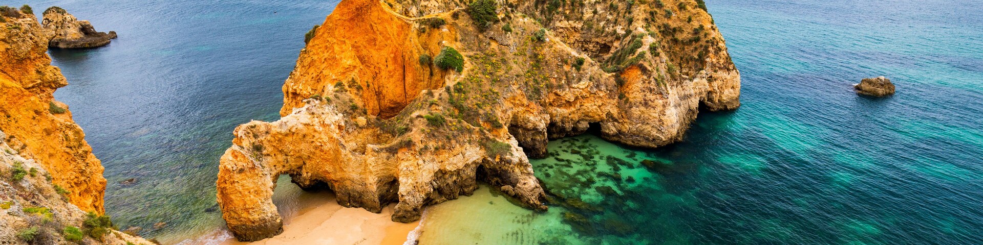 Paisagem costeira com uma formação rochosa imponente, águas turquesa cristalinas e uma praia de areia.