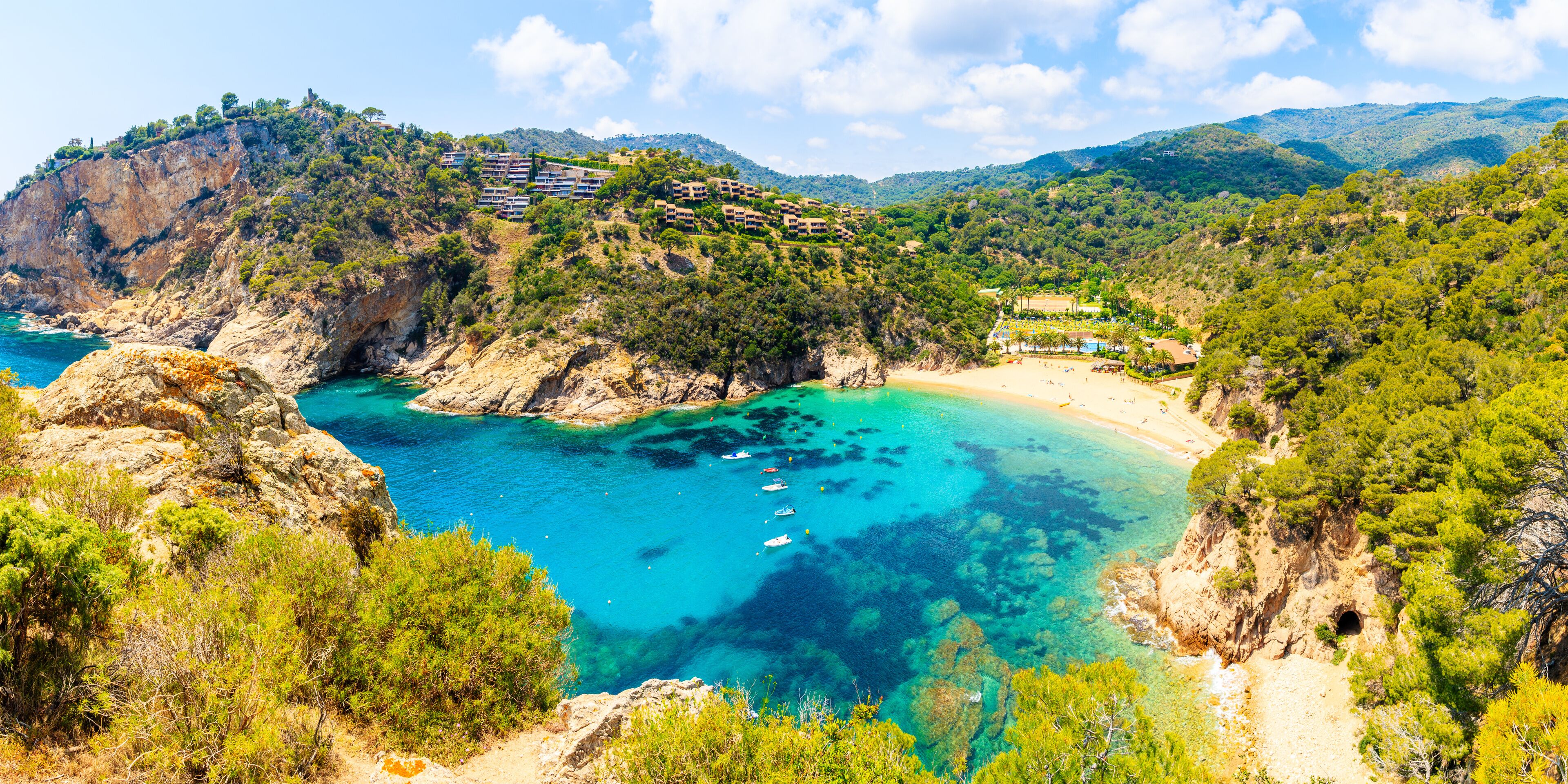 A coastal landscape with clear turquoise waters, a sandy beach, and a hillside with buildings.