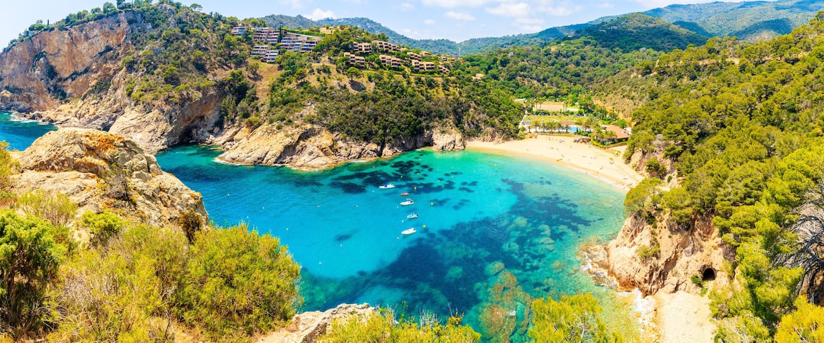 A coastal landscape with clear turquoise waters, a sandy beach, and a hillside with buildings.