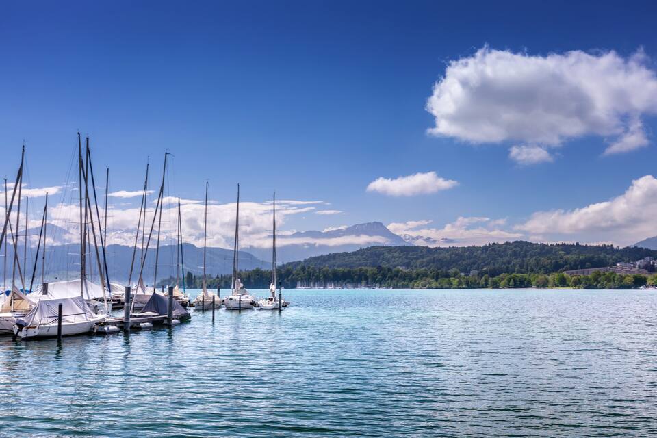 Un lago tranquilo con barcos a vela amarrados a la izquierda, rodeado de montañas y un cielo despejado.