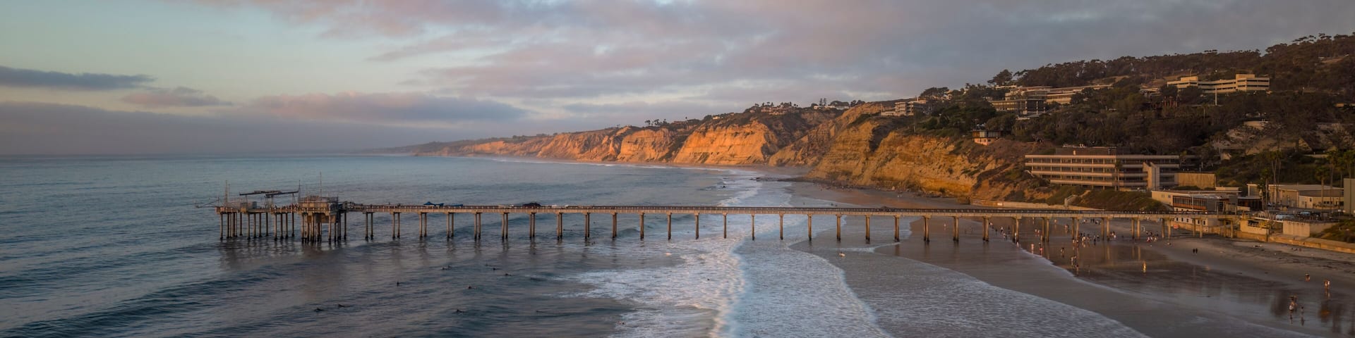 A coastal scene with a pier extending into the ocean, a sandy beach, and a cliff with buildings on top.