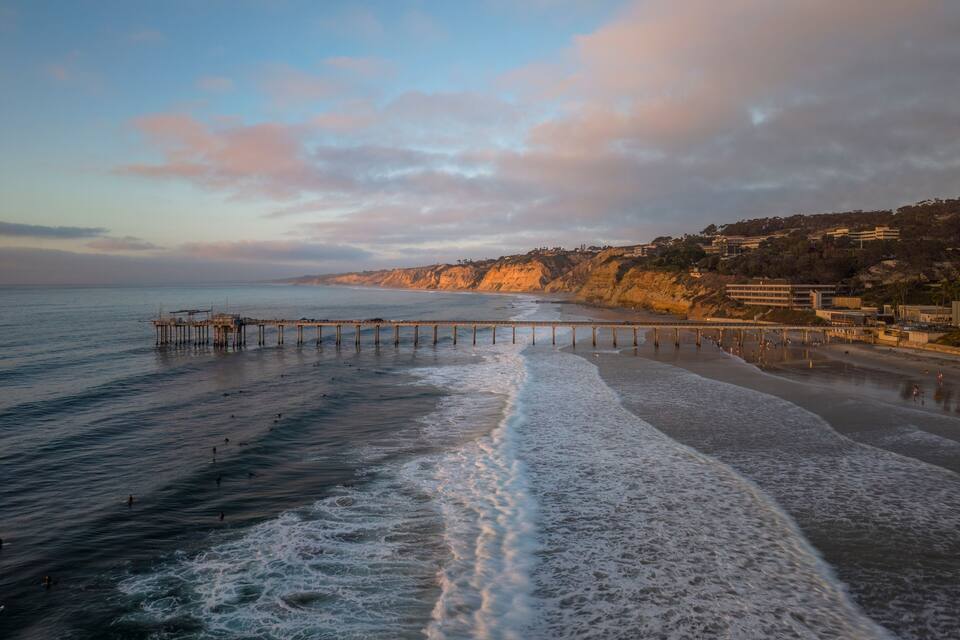 A coastal scene with a pier extending into the ocean, a sandy beach, and a cliff with buildings on top.