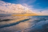 A sunset over the ocean with waves, a bird flying, and a partly cloudy sky.