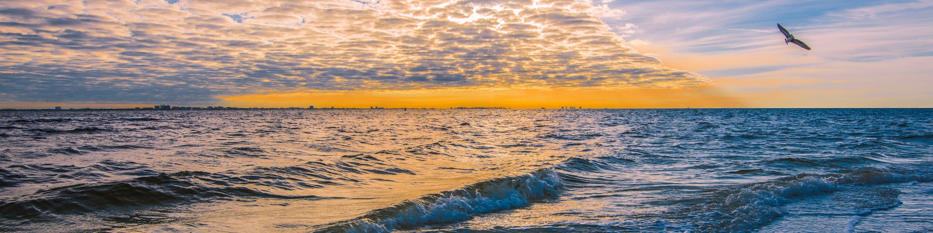 Pôr do sol sobre o oceano com ondas, um pássaro voando e céu parcialmente nublado.