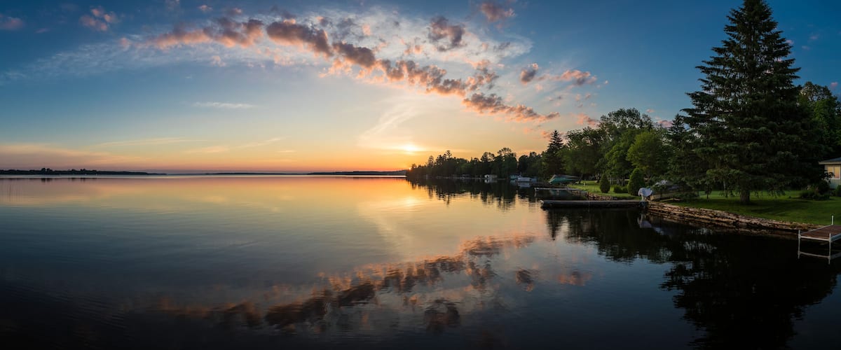 A calm lake at sunset with a dock and trees reflecting the sky.