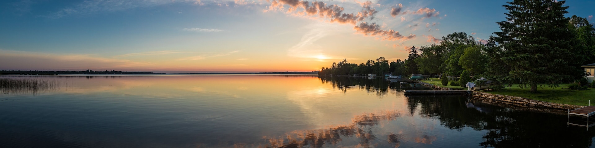 A calm lake at sunset with a dock and trees reflecting the sky.