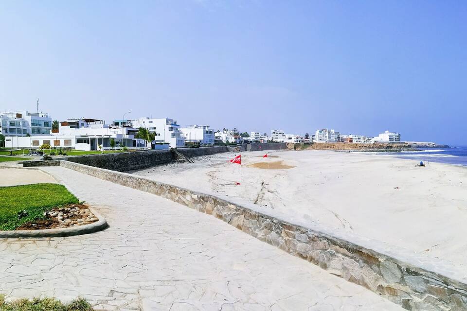 En kystpromenade med en stenvæg, en sandstrand og moderne hvide bygninger langs kysten.