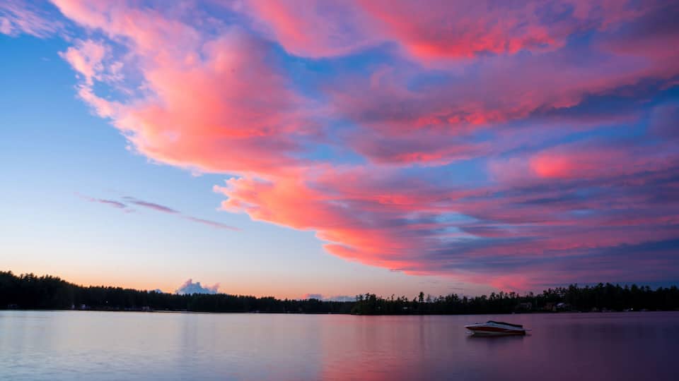 A calm lake with a boat, surrounded by a forest and a colorful sky.